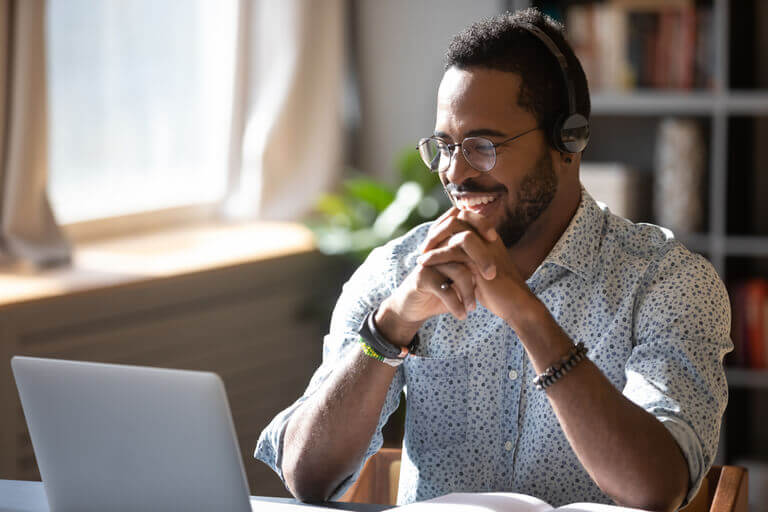 A smiling man wearing glasses and headphones sits at a desk, looking at a laptop. He appears to be in a video call or virtual meeting, with natural light coming through a window in the background. The setting looks like a home office or study.