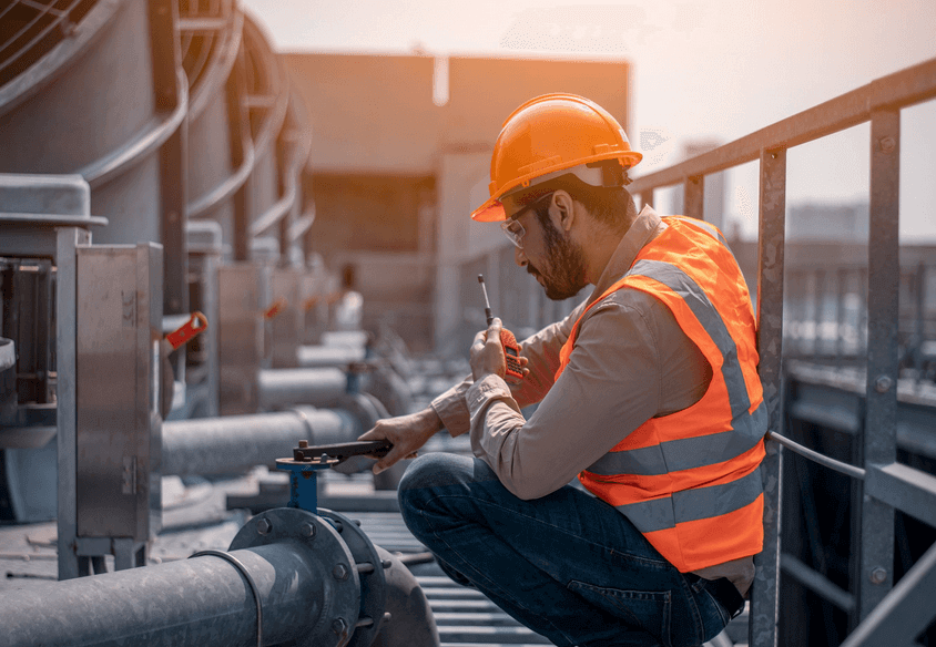 A man dressed in an orange vest and hard hat is sitting on a platform, engaged in his work environment.