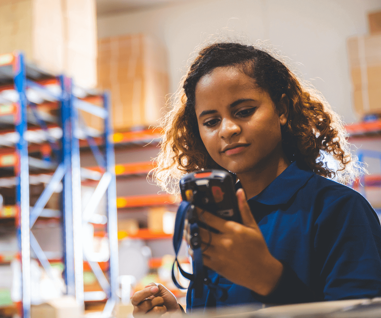 A young woman in a blue uniform uses a handheld barcode scanner in a warehouse with shelves of boxes in the background. The setting suggests inventory or logistics work.