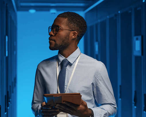 Man in a blue shirt, holding a tablet, stands in a dimly lit server room with blue lighting, examining equipment.