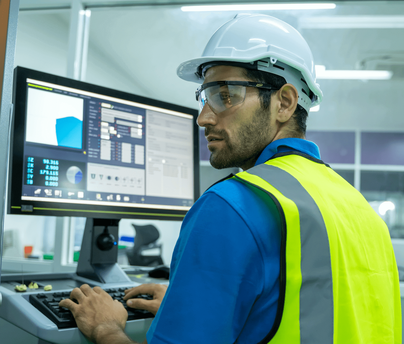 A man in a hard hat and safety vest is diligently operating a computer, emphasizing workplace safety and efficiency.